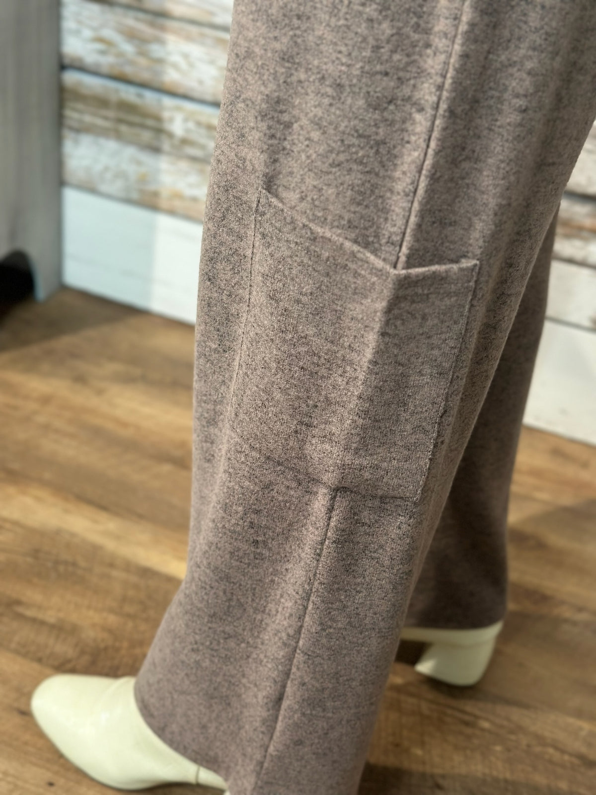 Close-up of a person wearing heather taupe wide-leg trousers with a side pocket, paired with white heeled boots, standing on a wooden floor.