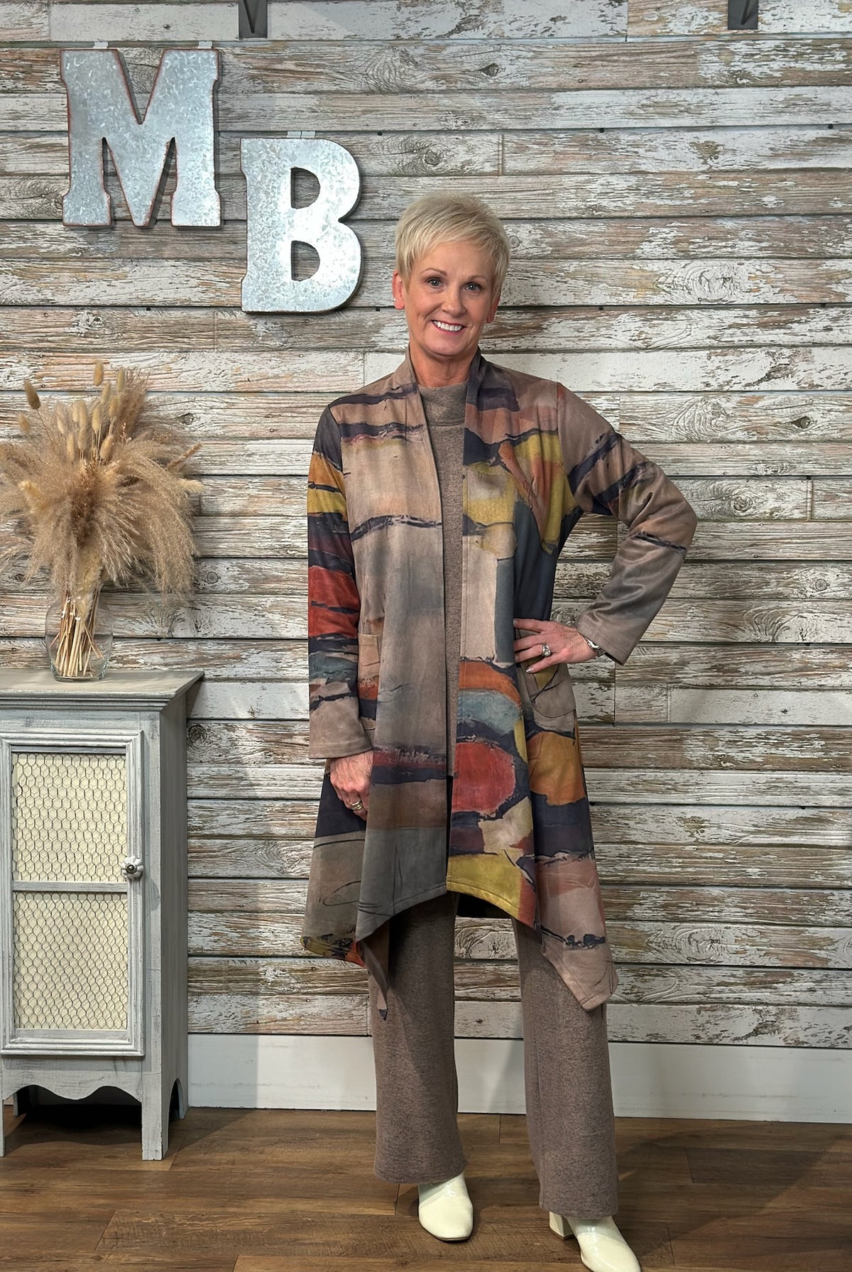 A woman modeling a long, open-front cardigan with an abstract multicolor pattern in earthy tones, paired with matching brown pants, standing in front of a rustic wood-paneled wall with decorative letters.