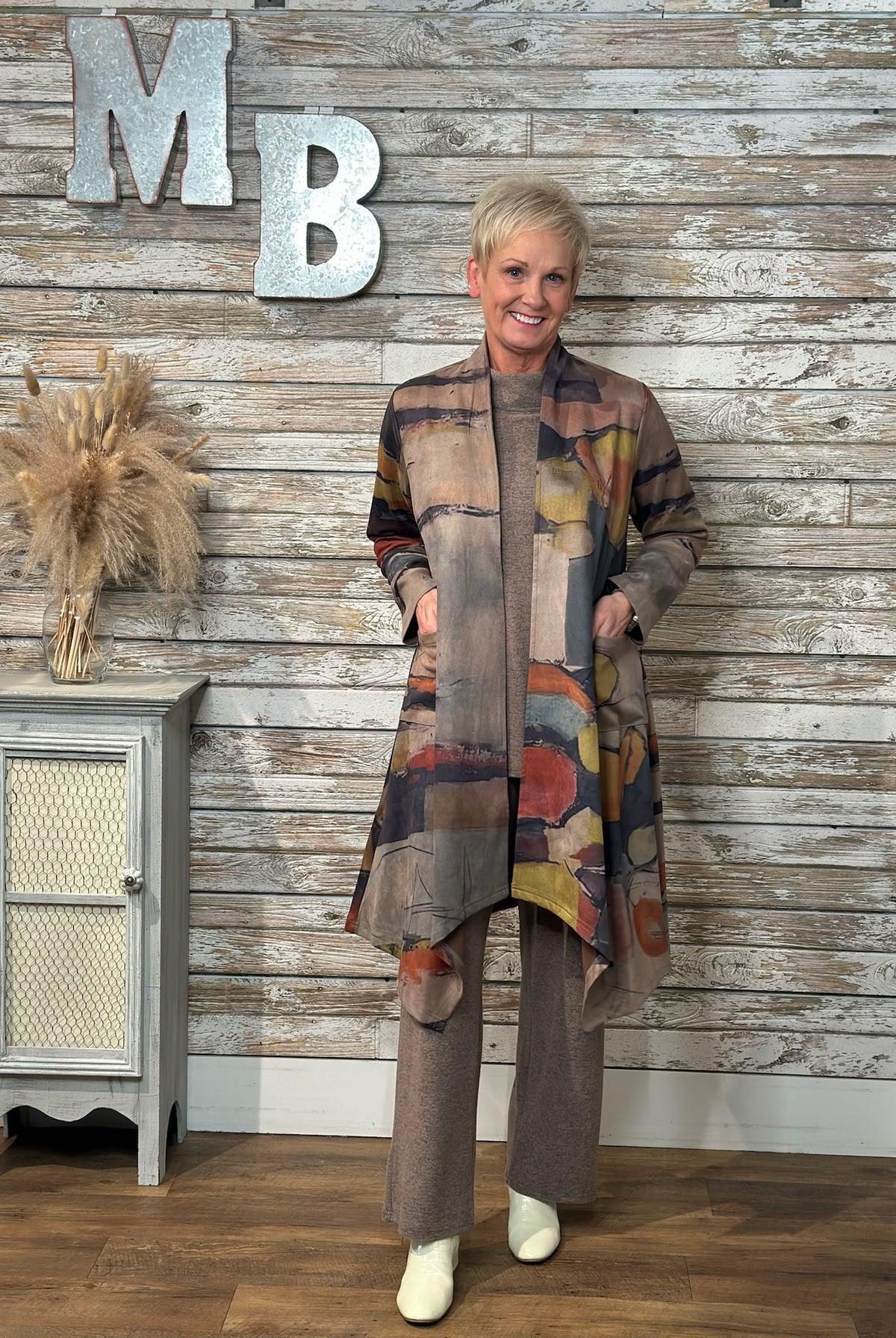 A woman stands in front of a rustic whitewashed wood wall wearing a long abstract-print cardigan over a taupe knit top and matching wide-leg pants, with white boots. Large metal letters 'MB' and a small cabinet with dried grass decor are visible in the background.