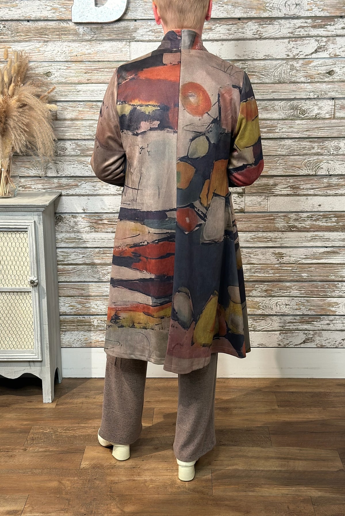 A person shown from the back wearing a long abstract-print coat with wide-leg taupe pants, standing in front of a rustic whitewashed wood wall and wood floor.