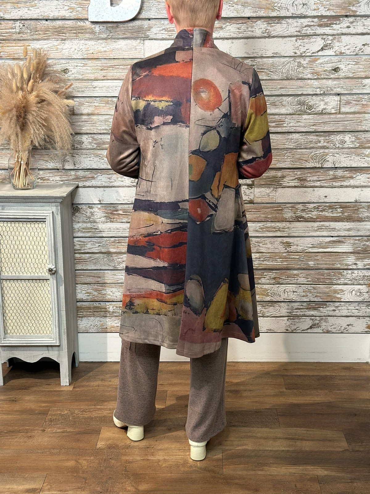 A person shown from the back wearing a long abstract-print coat with wide-leg taupe pants, standing in front of a rustic whitewashed wood wall and wood floor.