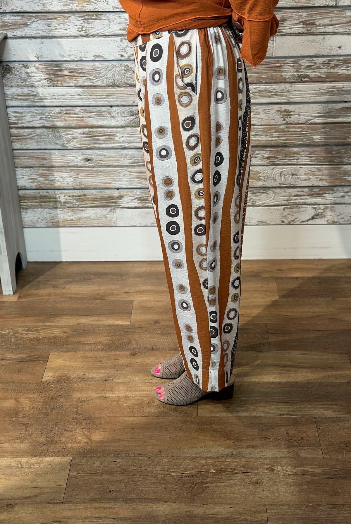 A person wearing loose, wide-leg pants with vertical rust-brown and white stripes, each printed with circular motifs in black, gray, and beige. The person is standing on a wood floor in front of a rustic whitewashed wood wall, wearing a rust-orange top and gray slip-on shoes.