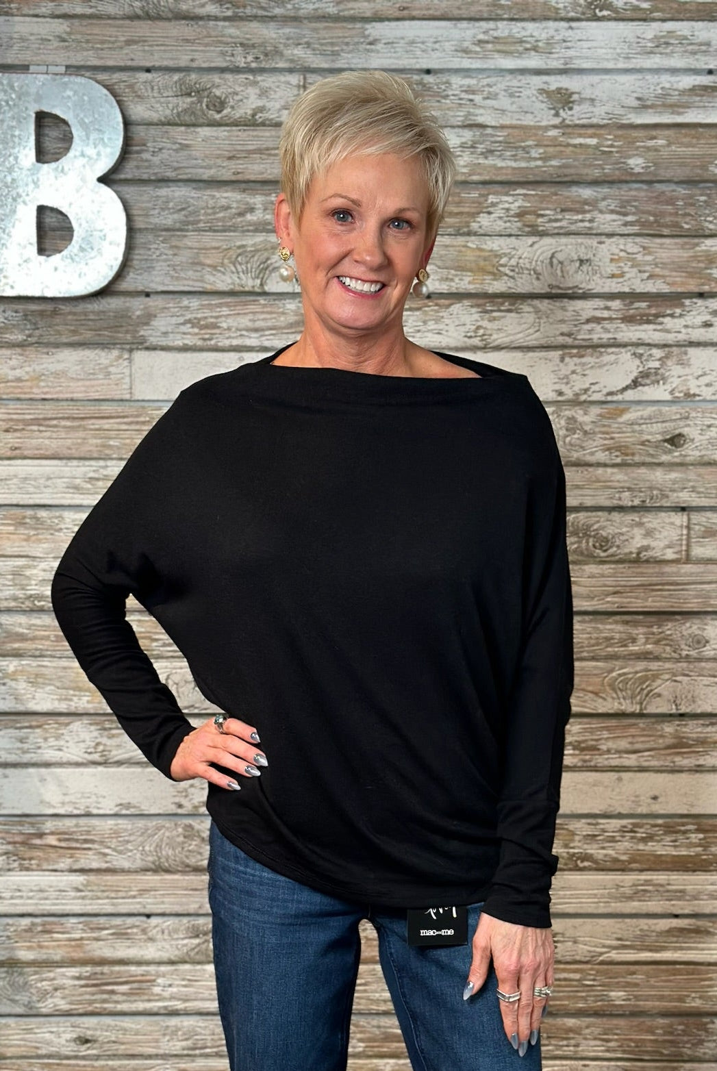A woman with short blonde hair wearing a black long-sleeve boat-neck top and blue jeans, standing in front of a rustic wood plank wall with a large silver letter 'B' on the left.