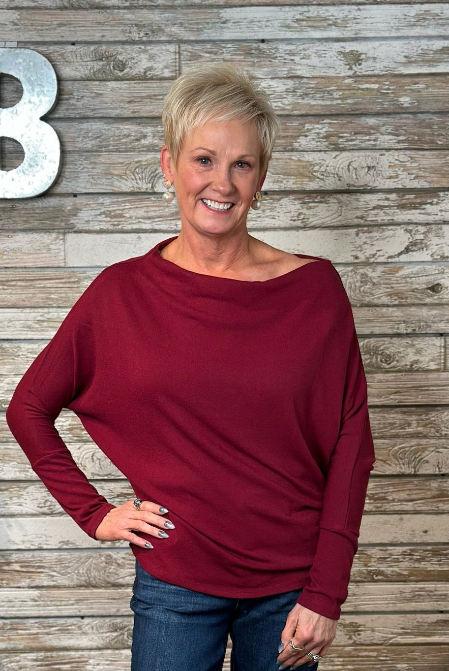 A smiling blonde woman wearing a long-sleeve, boat-neck, slouchy knit top in deep burgundy, paired with blue jeans, standing against a rustic gray wood plank wall with a large silver letter 'B' on the left.