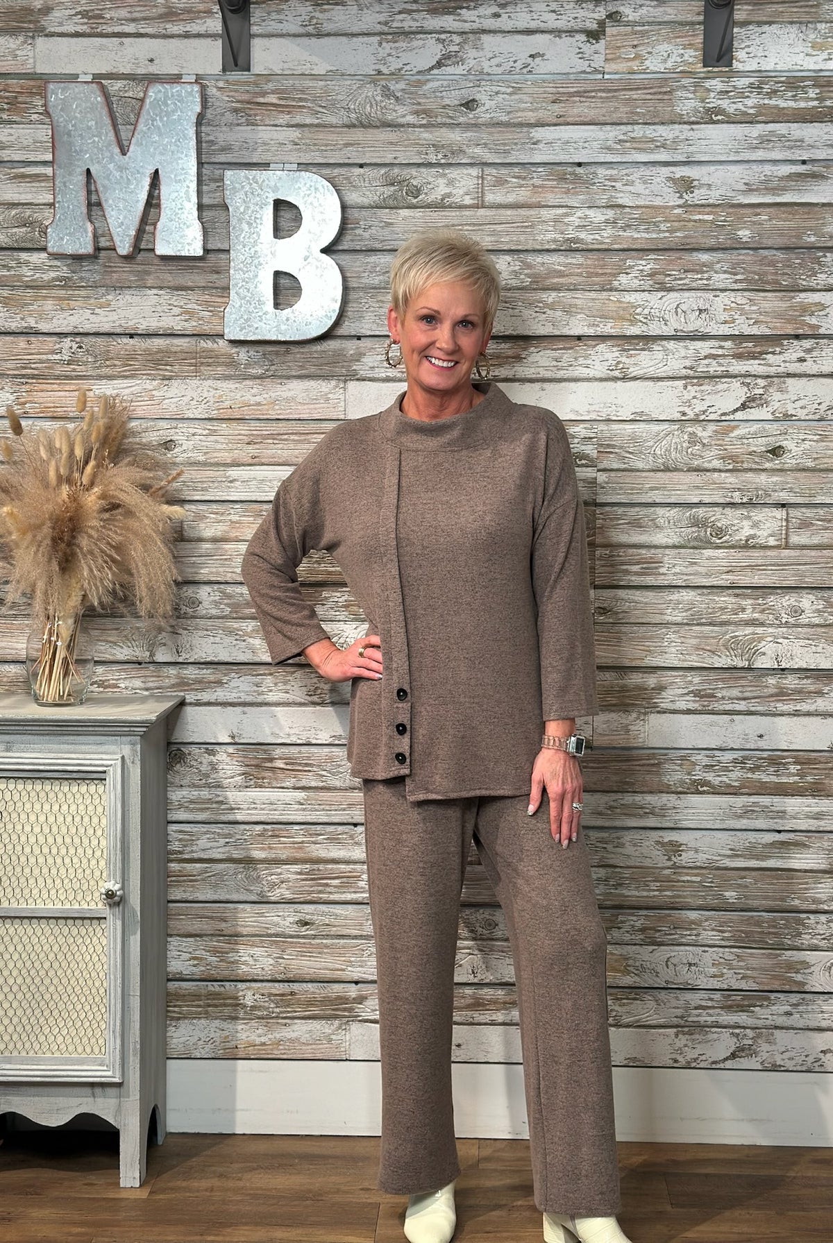 A woman modeling a matching taupe brown knit two-piece set consisting of a long-sleeve button-front top and wide-leg pants, standing in front of a rustic whitewashed wood plank wall with decorative 'MB' letters.