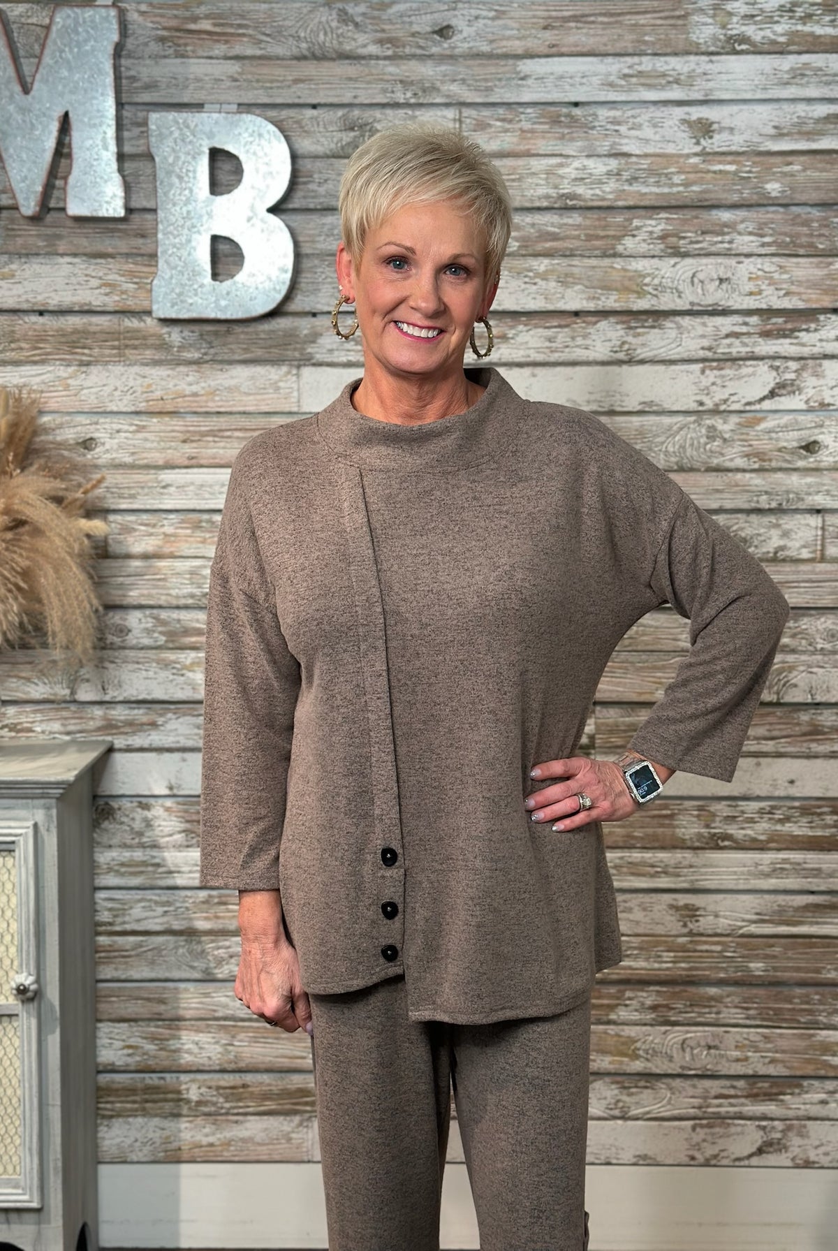 A woman wearing a matching taupe-brown knit top and pants set with black buttons on the front of the top. Large decorative letters 'MB' are visible on the wall, and a small cabinet with dried grass decor appears on the left.