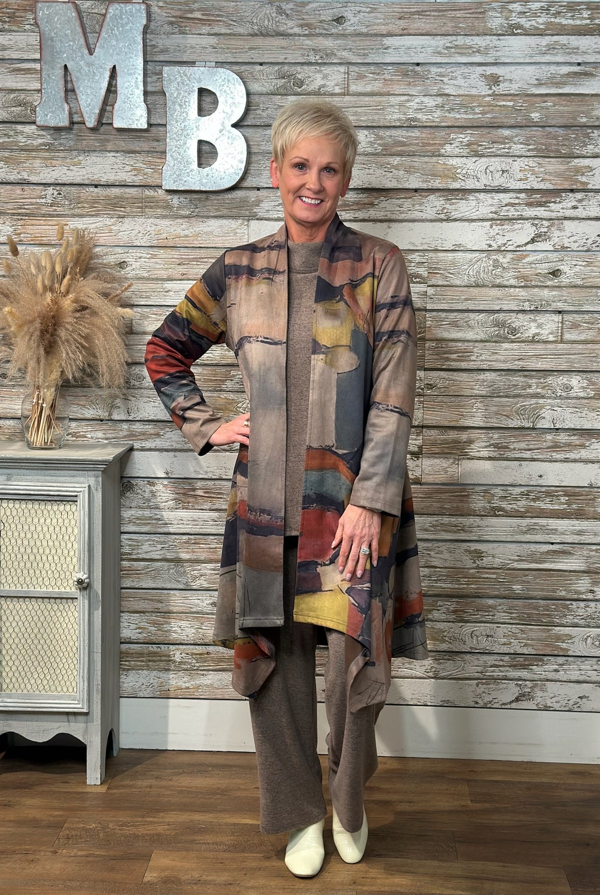 A woman wearing a long patchwork cardigan with abstract multicolor blocks in beige, brown, gray, blue, and rust tones, paired with wide-leg taupe pants and white shoes.