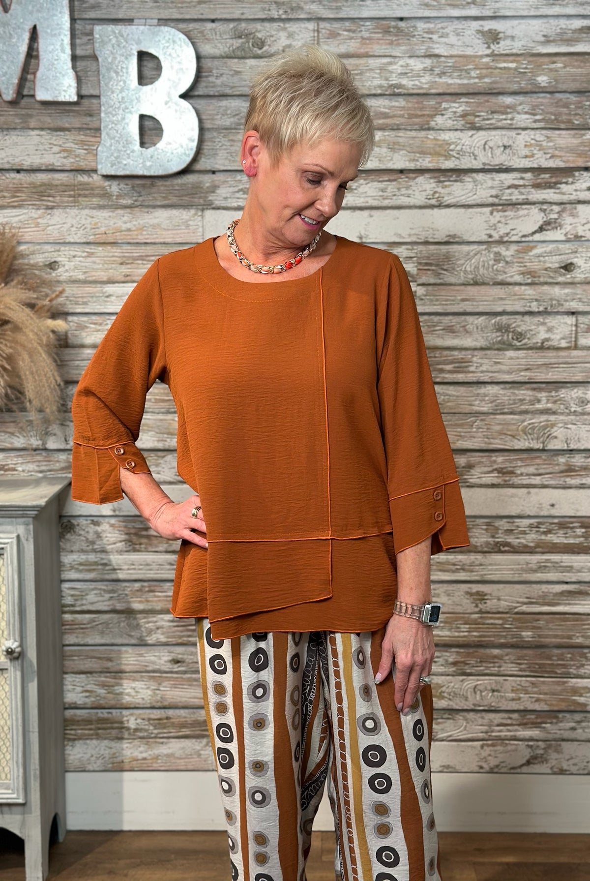 A woman modeling a rust-orange textured three-quarter sleeve top with button details on the cuffs, paired with patterned pants, standing against a rustic wood-paneled wall.