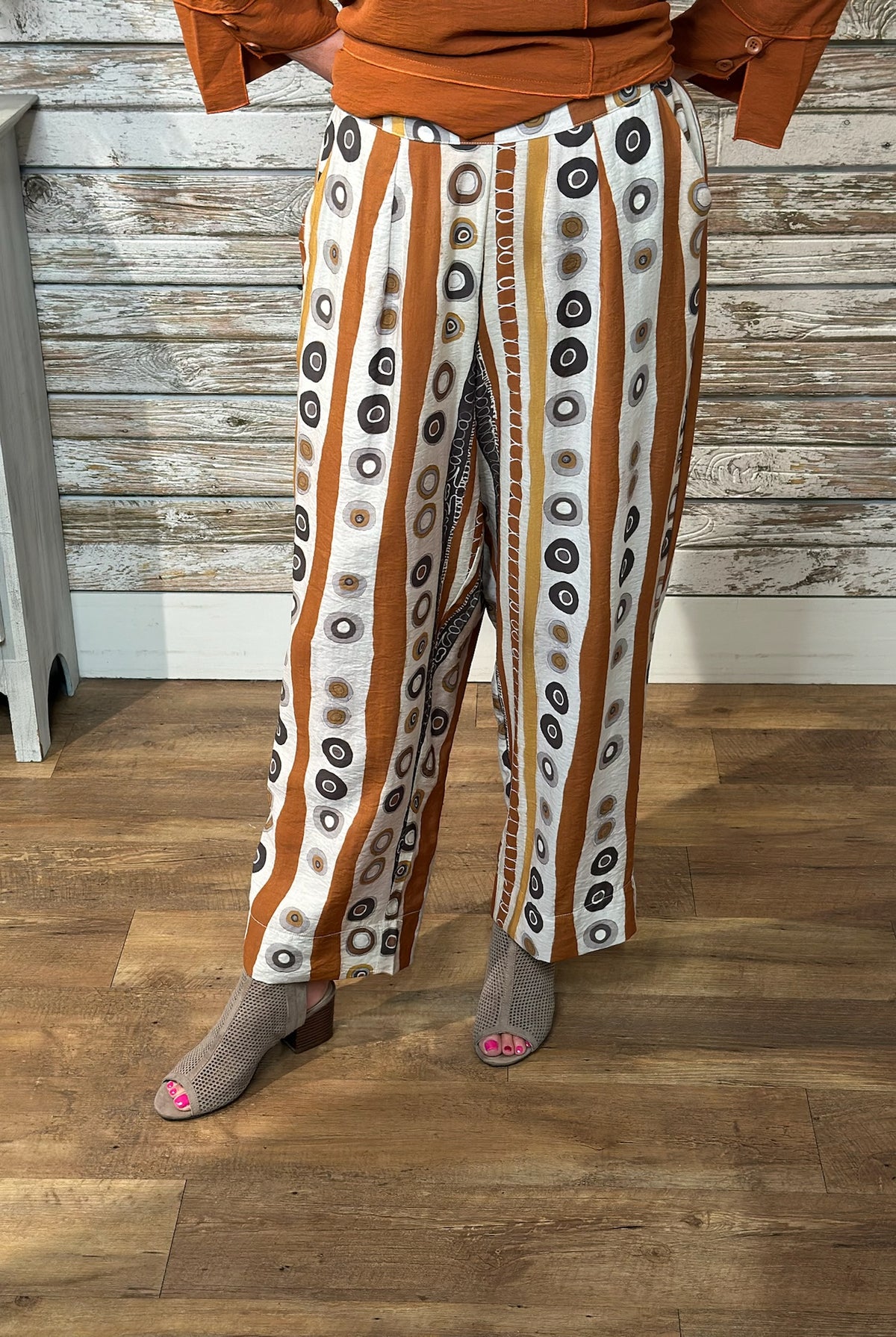 A person wearing wide-leg cropped pants with vertical rust-orange and white stripes and circular black, gray, and brown motifs, paired with an orange top and gray peep-toe heels, standing on a wood floor against a rustic wood plank wall.