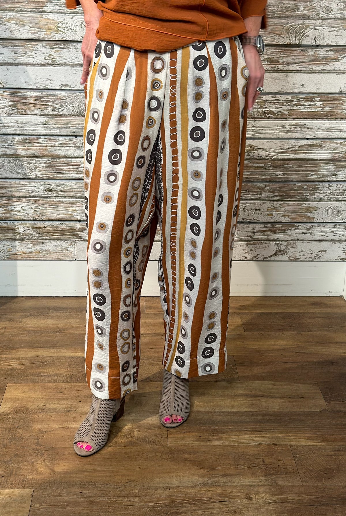 A person wearing wide-leg cropped pants with vertical stripes of brown, white, and black circular motifs, paired with an orange-brown top and gray slip-on shoes, standing against a rustic wood plank wall and floor.
