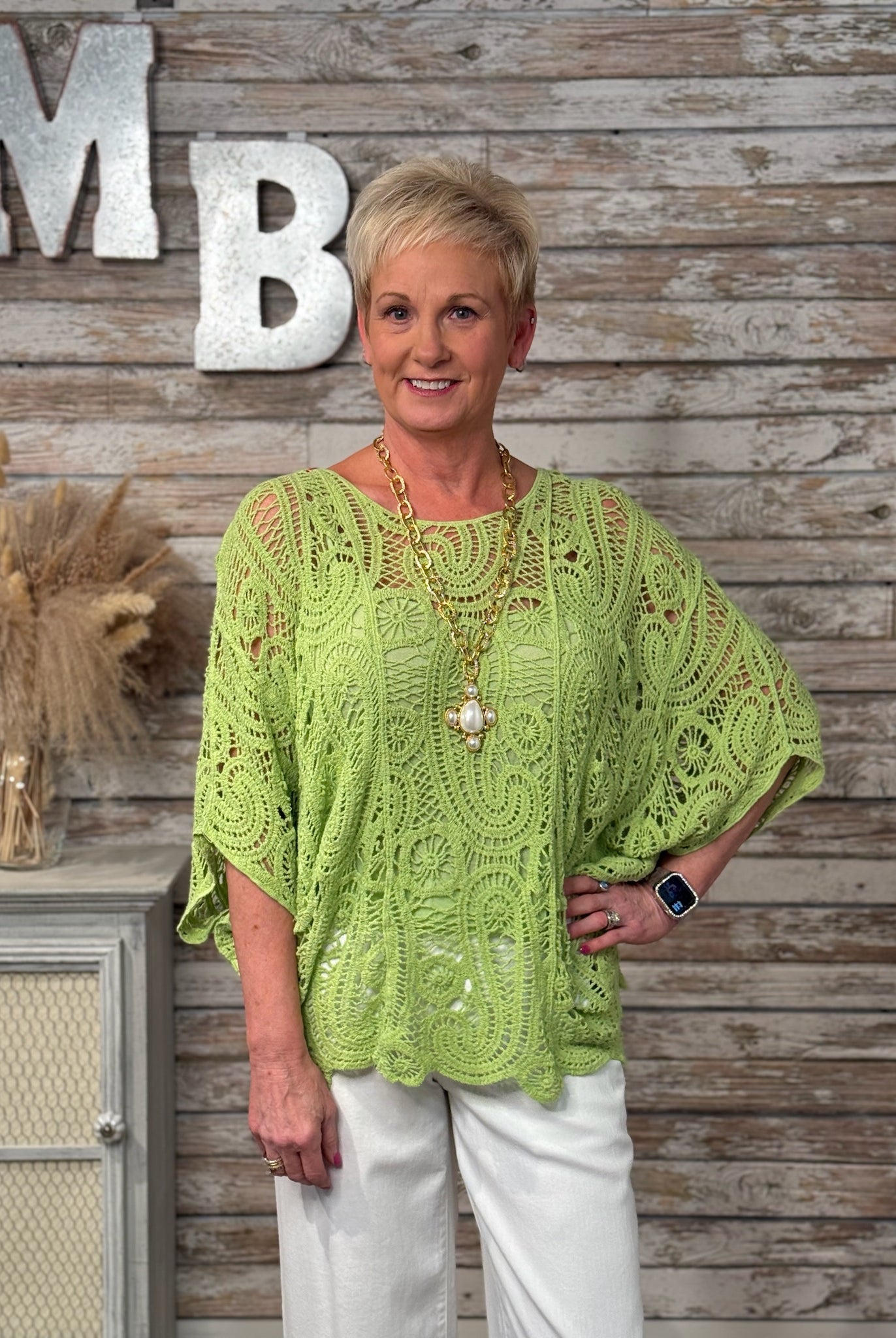 Woman wearing a green lace top standing in front of a wooden wall with decorative letters.