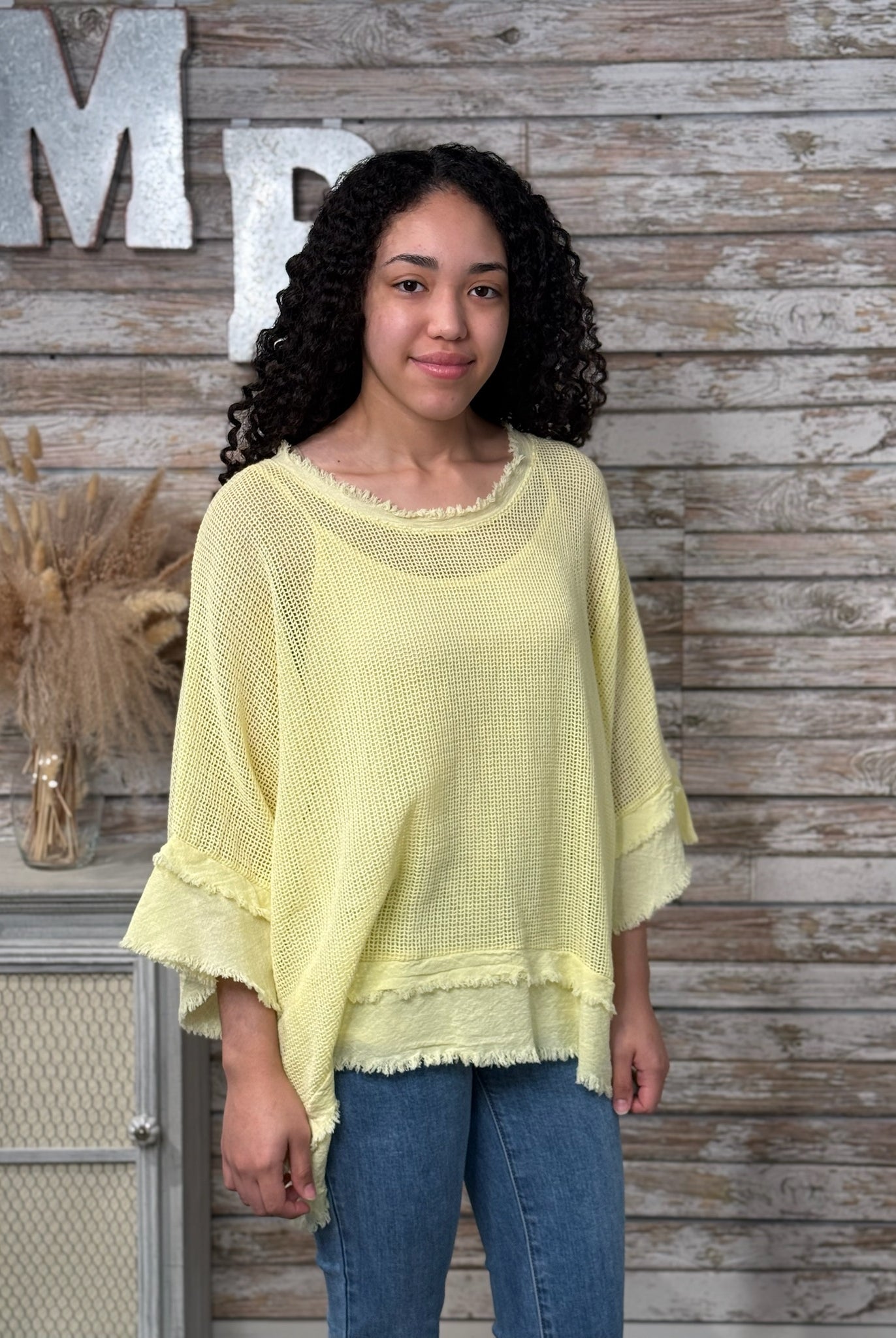 Woman wearing a light yellow top and blue jeans standing against a wooden wall with decorative letters.