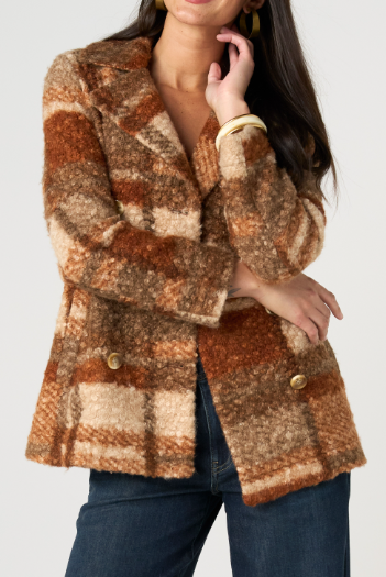 A woman wearing a fuzzy, textured plaid blazer in warm brown, beige, and rust tones, paired with dark blue jeans, against a solid light gray background.