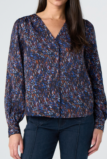 A woman wearing a long-sleeve V-neck blouse with a blue, brown, and white abstract speckled pattern, paired with dark blue jeans, against a light gray studio background.
