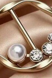 A gold-tone circular brooch or pin featuring a single pearl and three clear rhinestones, displayed on a soft beige satin fabric.