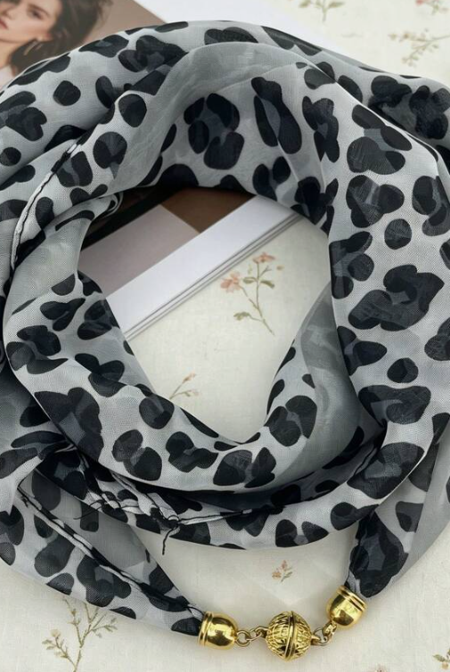 A lightweight scarf with a black leopard print on a light gray background, styled in a loop with gold-toned decorative beads at the end, resting on a floral fabric surface.