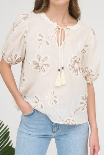 Woman wearing a beige embroidered blouse with puff sleeves and blue jeans against a white background.