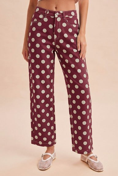 A model wearing high-waisted, wide-leg trousers in a deep burgundy color with a white polka dot pattern, paired with white espadrille flats, set against a solid light beige background.