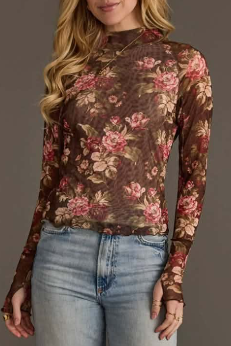 A woman wearing a sheer long-sleeve mock-neck top with a brown floral print in pink and beige, paired with light-wash blue jeans, against a solid dark gray background.