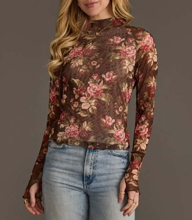 A woman wearing a sheer long-sleeve mock-neck top with a brown floral print in pink and beige, paired with light-wash blue jeans, against a solid dark gray background.