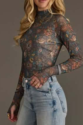 A woman wearing a sheer long-sleeve mock-neck top with an intricate floral and paisley pattern in muted earthy tones, paired with light-wash blue jeans, set against a solid dark gray studio background.