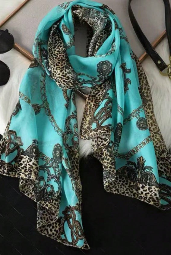 A turquoise scarf with a leopard print border and a black chain-link pattern, styled on a white faux fur surface with sunglasses and a handbag nearby.