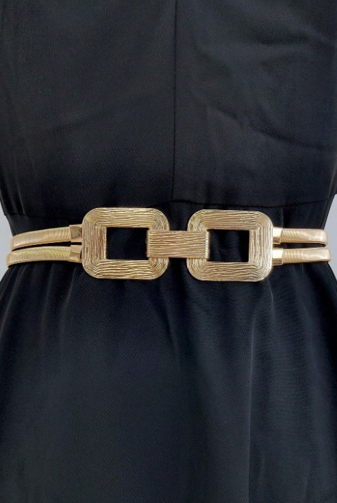 A gold-colored belt with a textured finish and a large rectangular link design, worn as a waist accessory over a black dress.