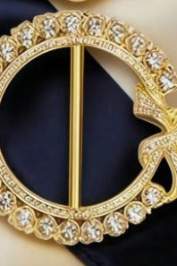 A gold-tone circular brooch with a decorative bow at the right side, set with clear rhinestones, displayed on a dark navy blue fabric background.