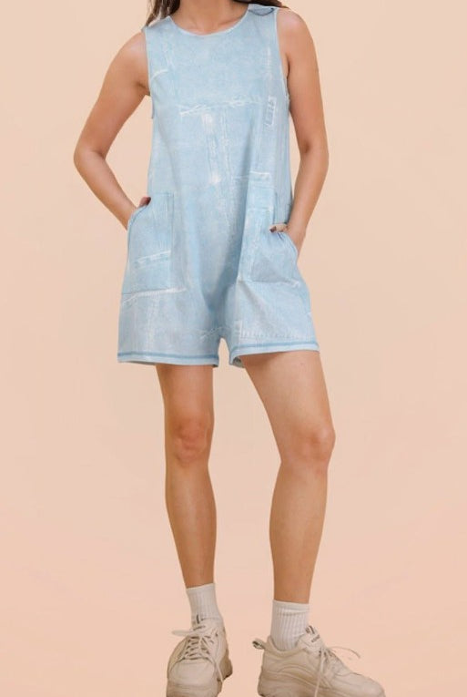 A model wearing a light blue denim romper with front patch pockets, paired with white socks and beige sneakers, set against a solid pale peach background.