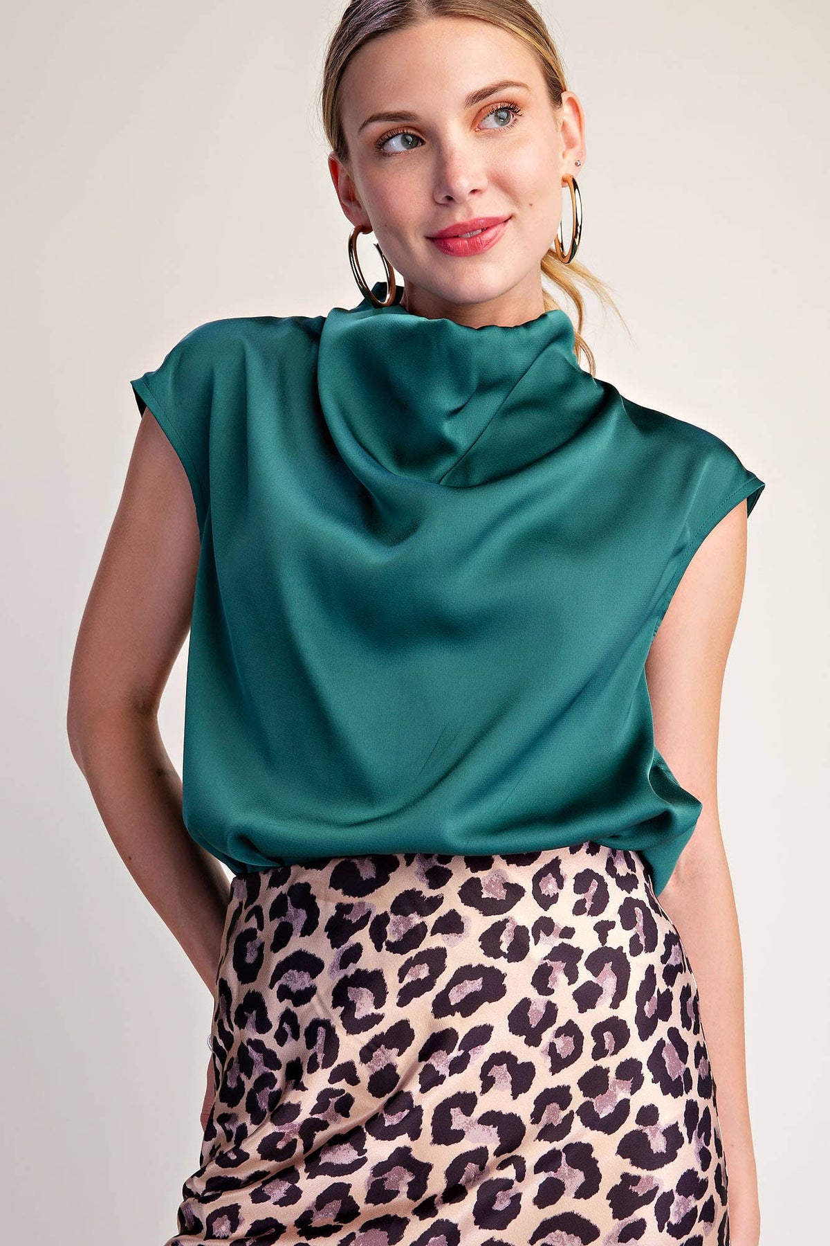 A woman wearing a teal green satin cowl-neck top and a leopard-print skirt, photographed against a plain light beige studio background.