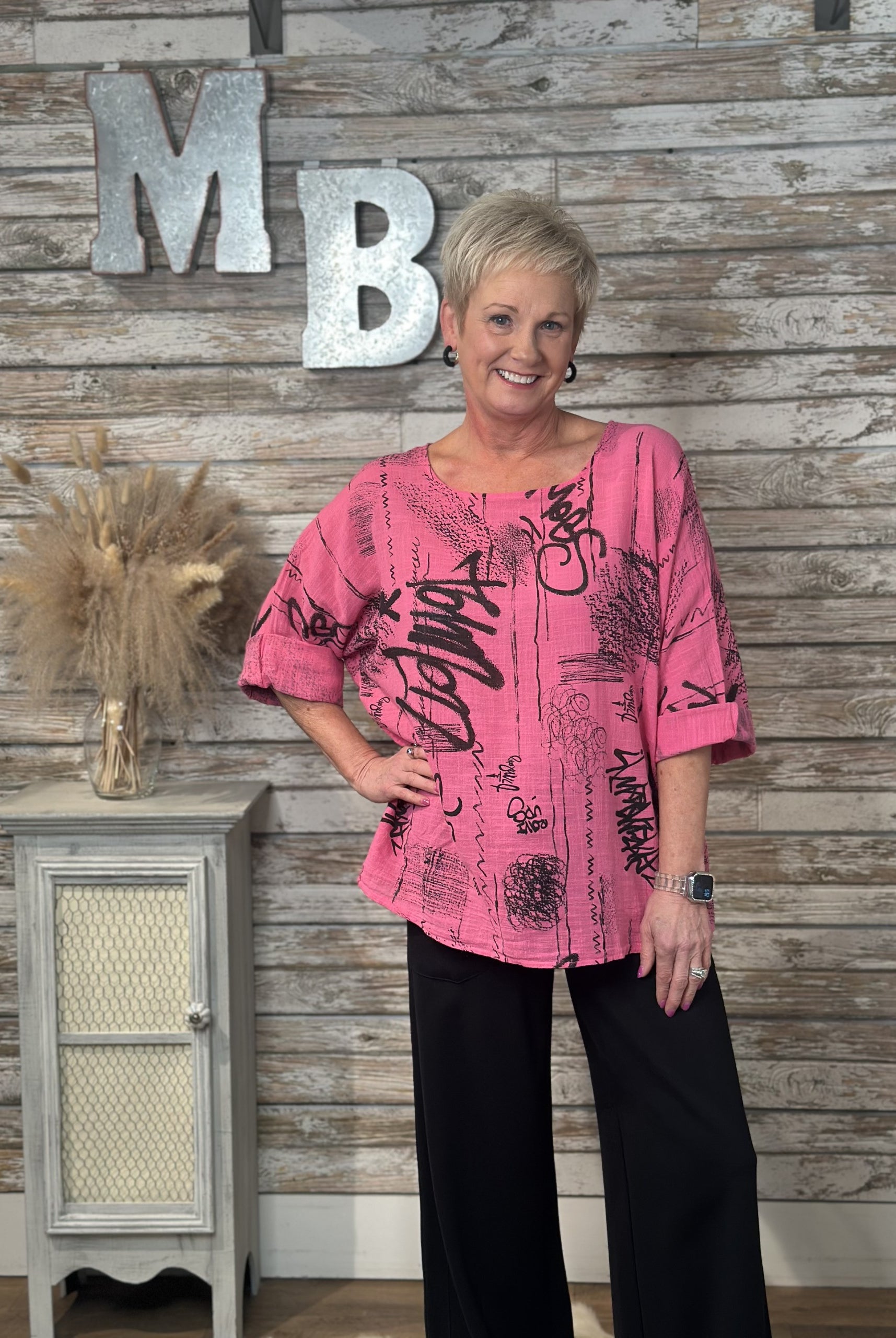 Woman in a pink top with black patterns standing in front of a wooden wall with 'MB' letters.