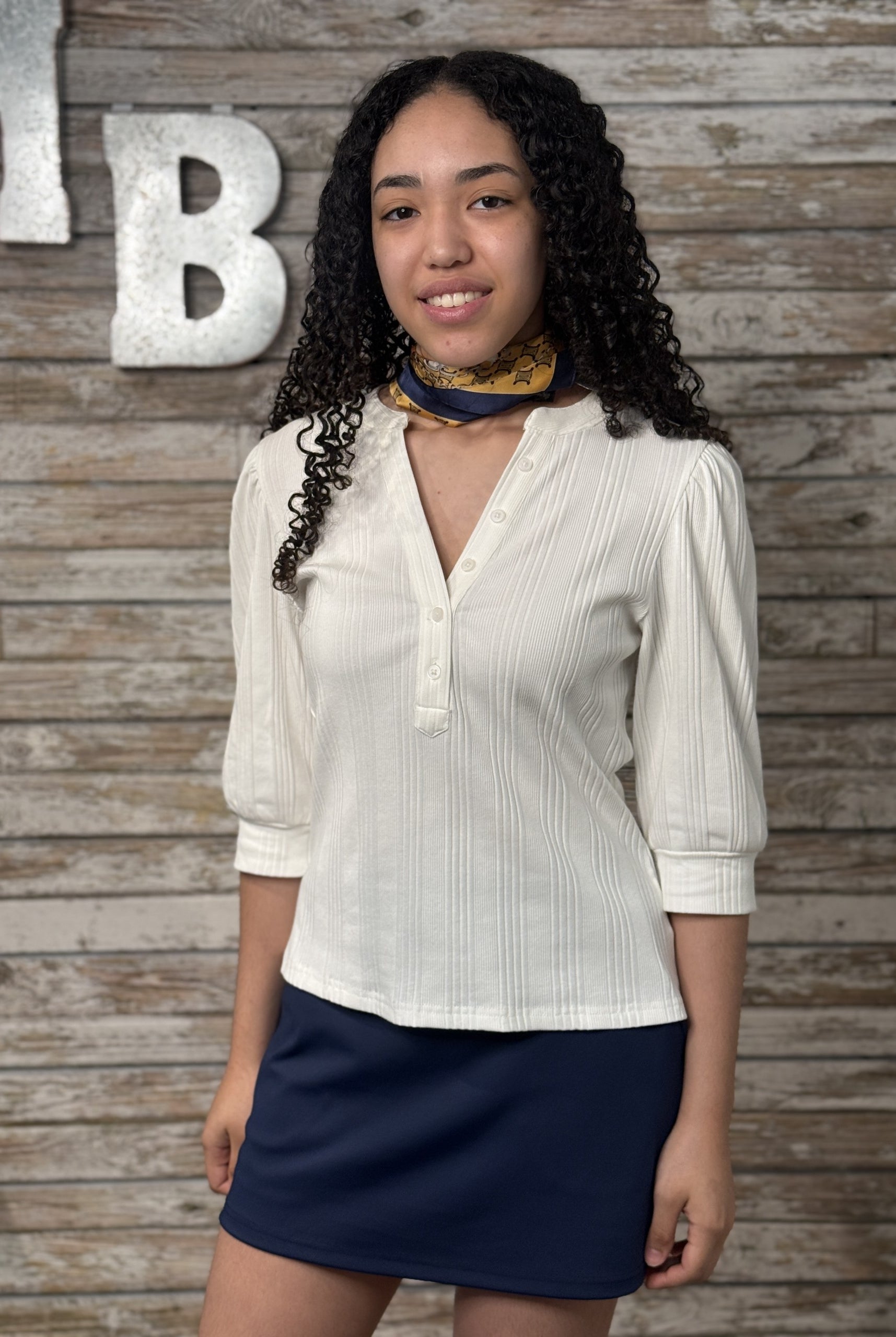 Woman wearing a white blouse and navy skirt against a wooden wall with decorative letters.