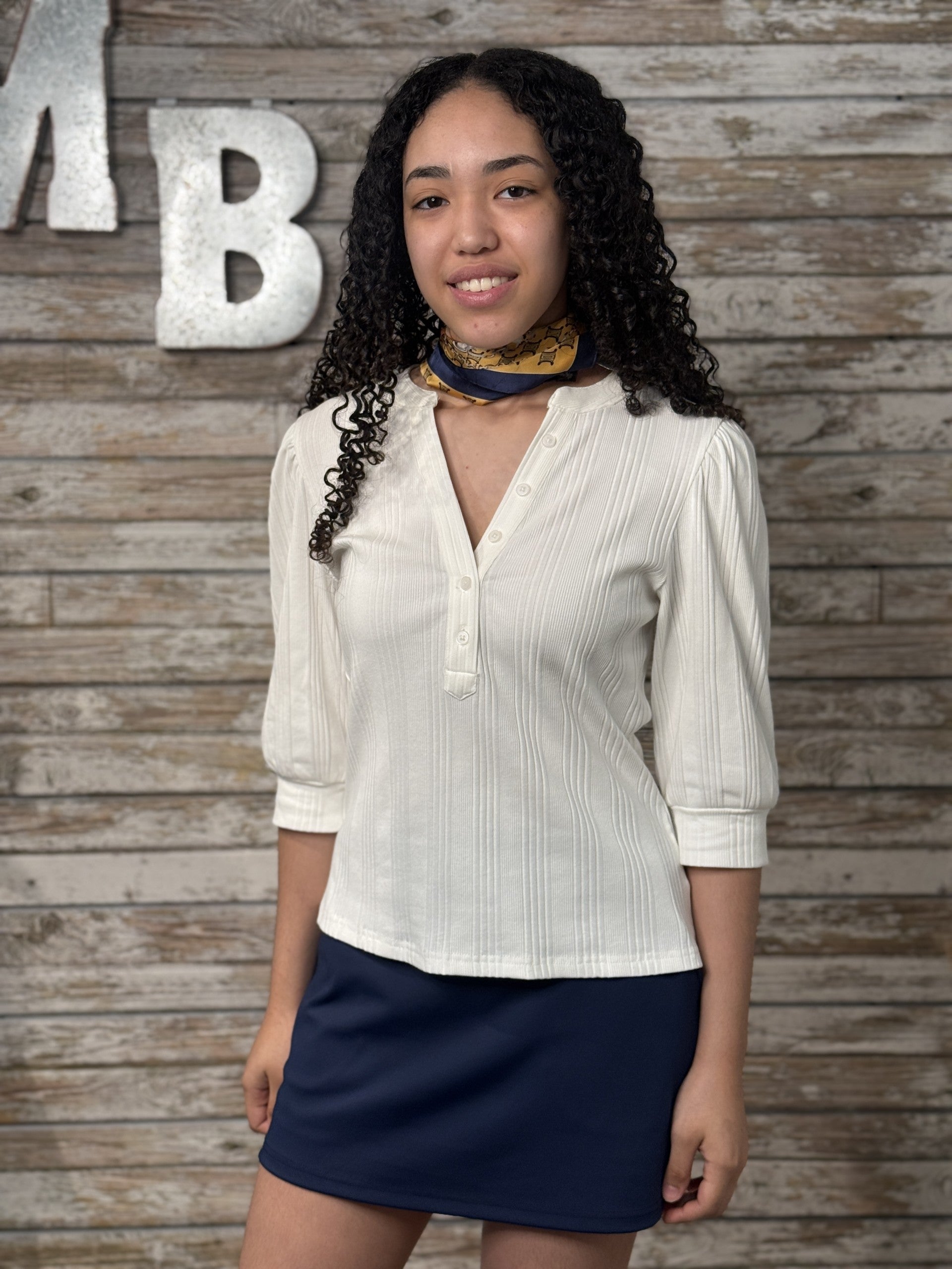 Woman wearing a white blouse and navy skirt against a wooden wall with decorative letters.