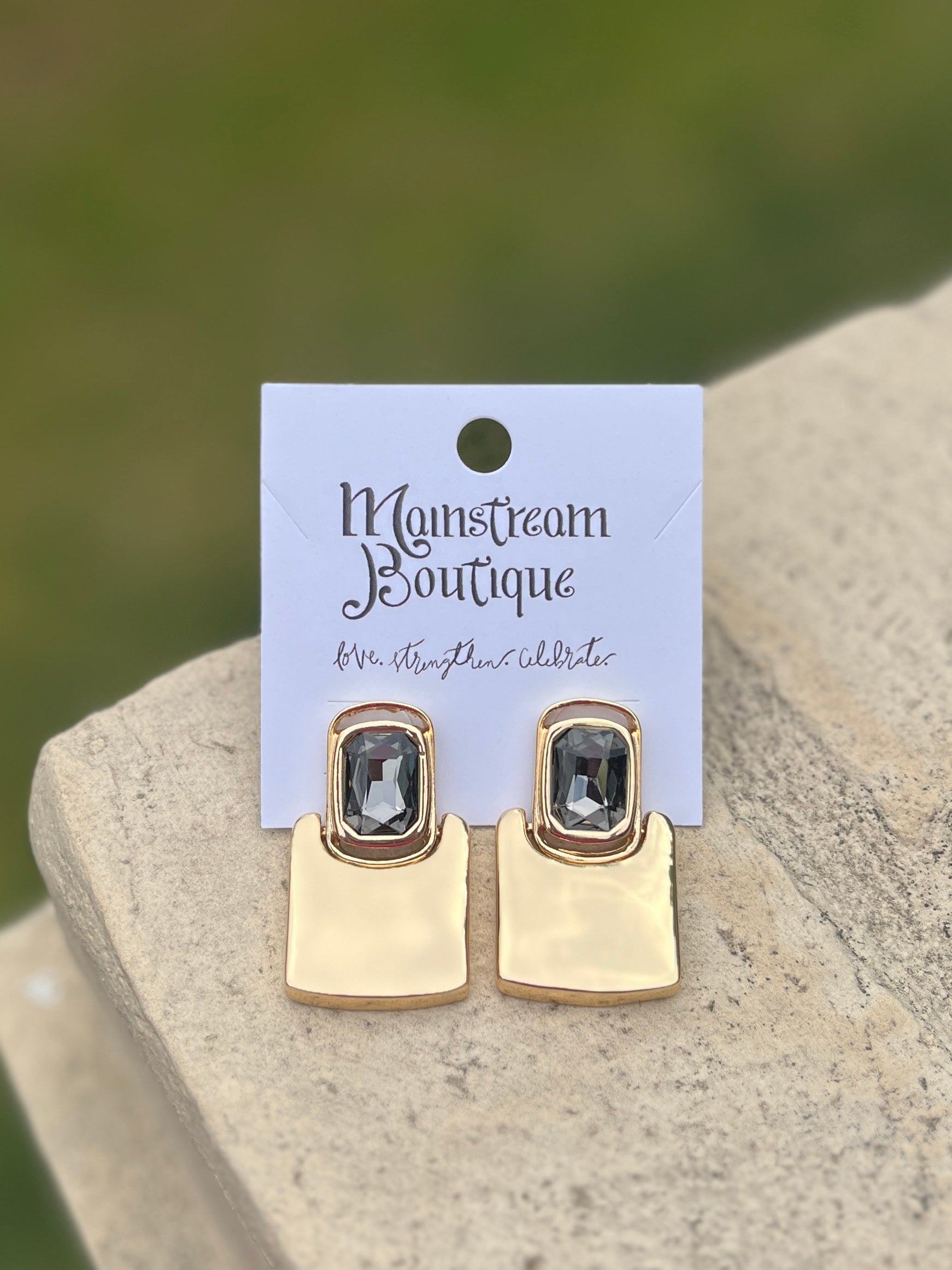 Gold earrings with gemstones placed on a stone surface with a blurred green background.