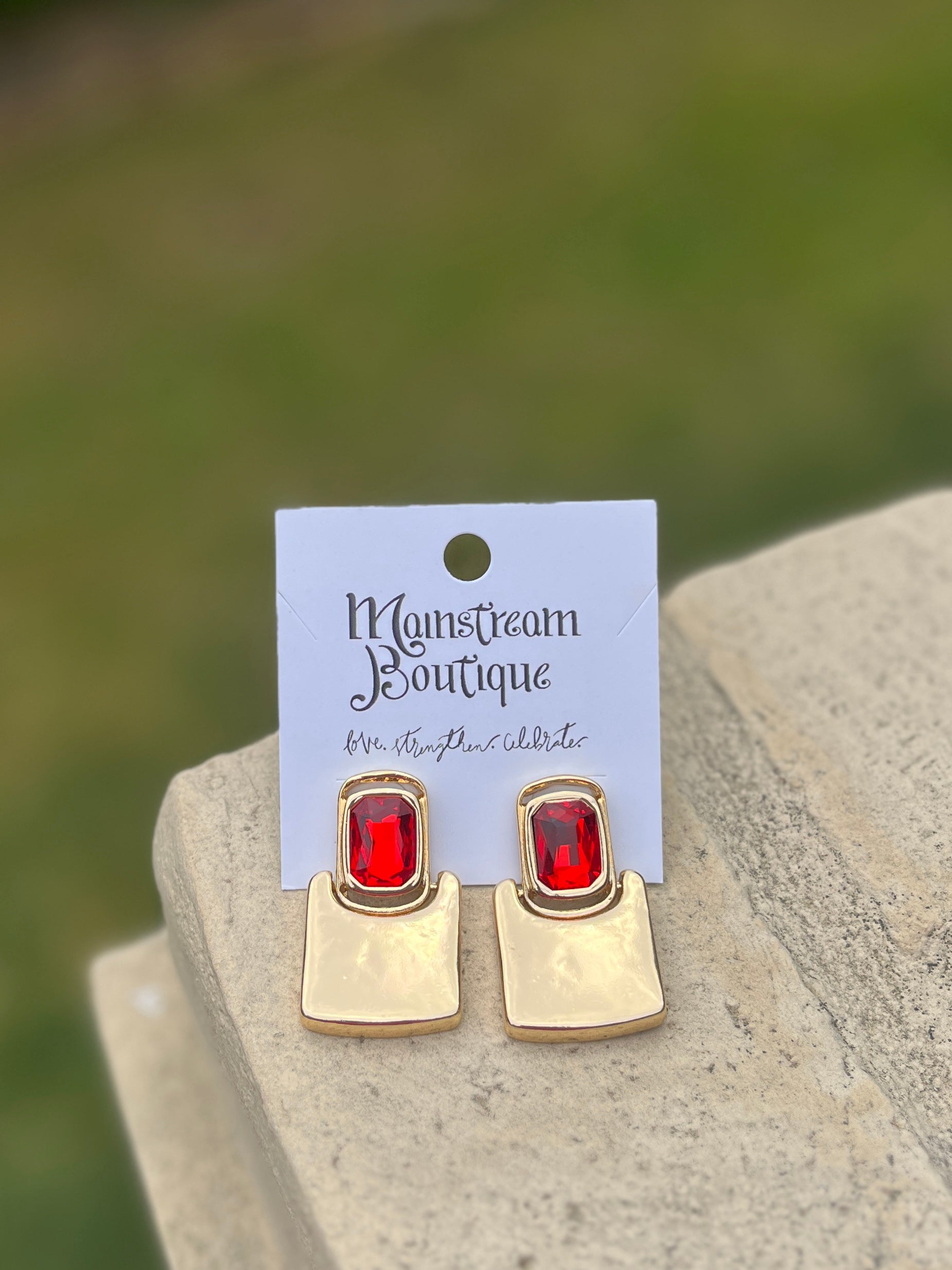 Gold earrings with red stones against a blurred green background.