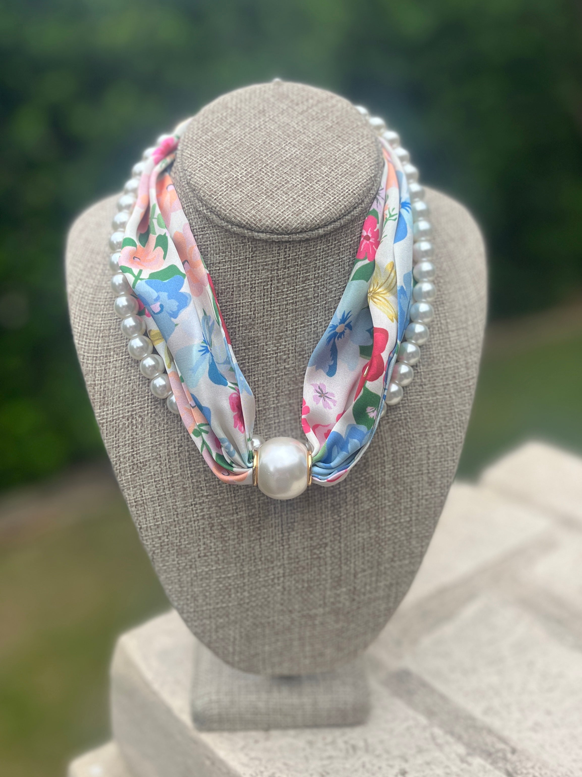 Necklace scarf with floral ribbon and pearls on a gray bust against a blurred green background