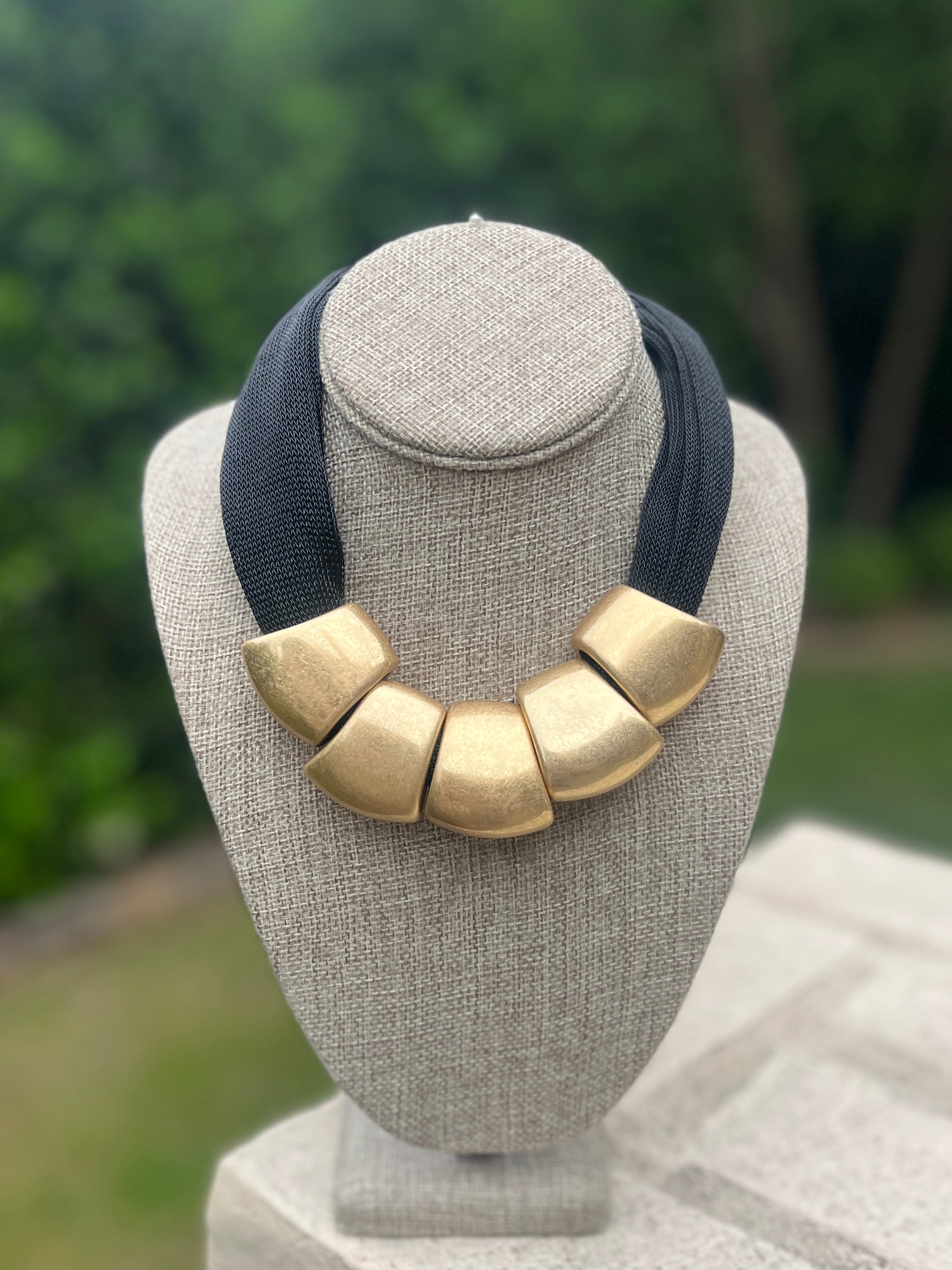 Necklace with gold bibs and navy strap on a gray mannequin bust with a blurred green background