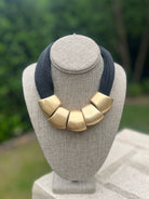Necklace with gold bibs and navy strap on a gray mannequin bust with a blurred green background
