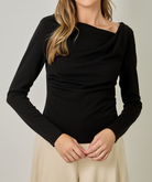 A woman wearing a fitted black long-sleeve top with an asymmetrical draped neckline and cream-colored pants, standing against a plain light gray studio background.