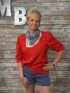 Woman wearing a red blouse and blue shorts standing against a wooden wall with decorative letters.