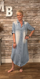 A woman modeling a light blue denim button-down shirt dress with a faded white wash effect, standing in front of a rustic wood-paneled wall.
