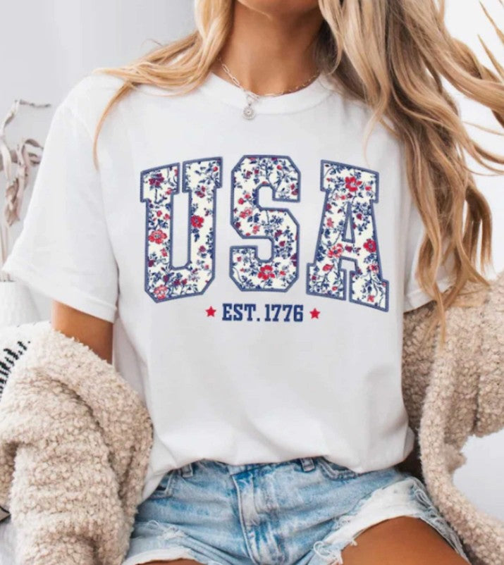 Person wearing a white t-shirt with 'USA' and floral design, sitting on a couch.