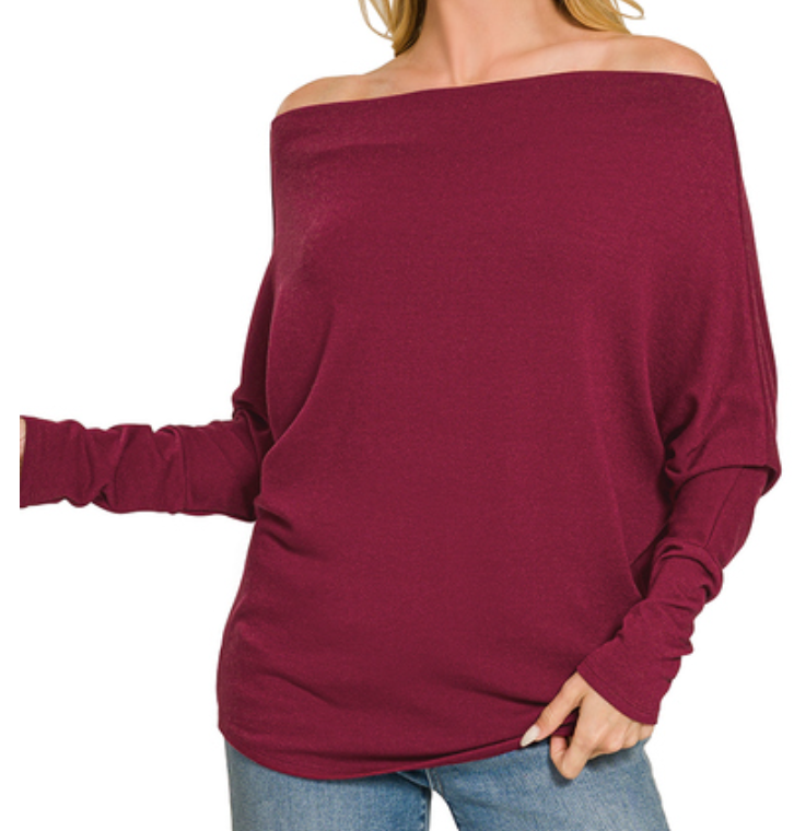 A woman wearing a burgundy off-the-shoulder long-sleeve knit top with gathered sleeves, paired with blue jeans, shown against a plain white background.
