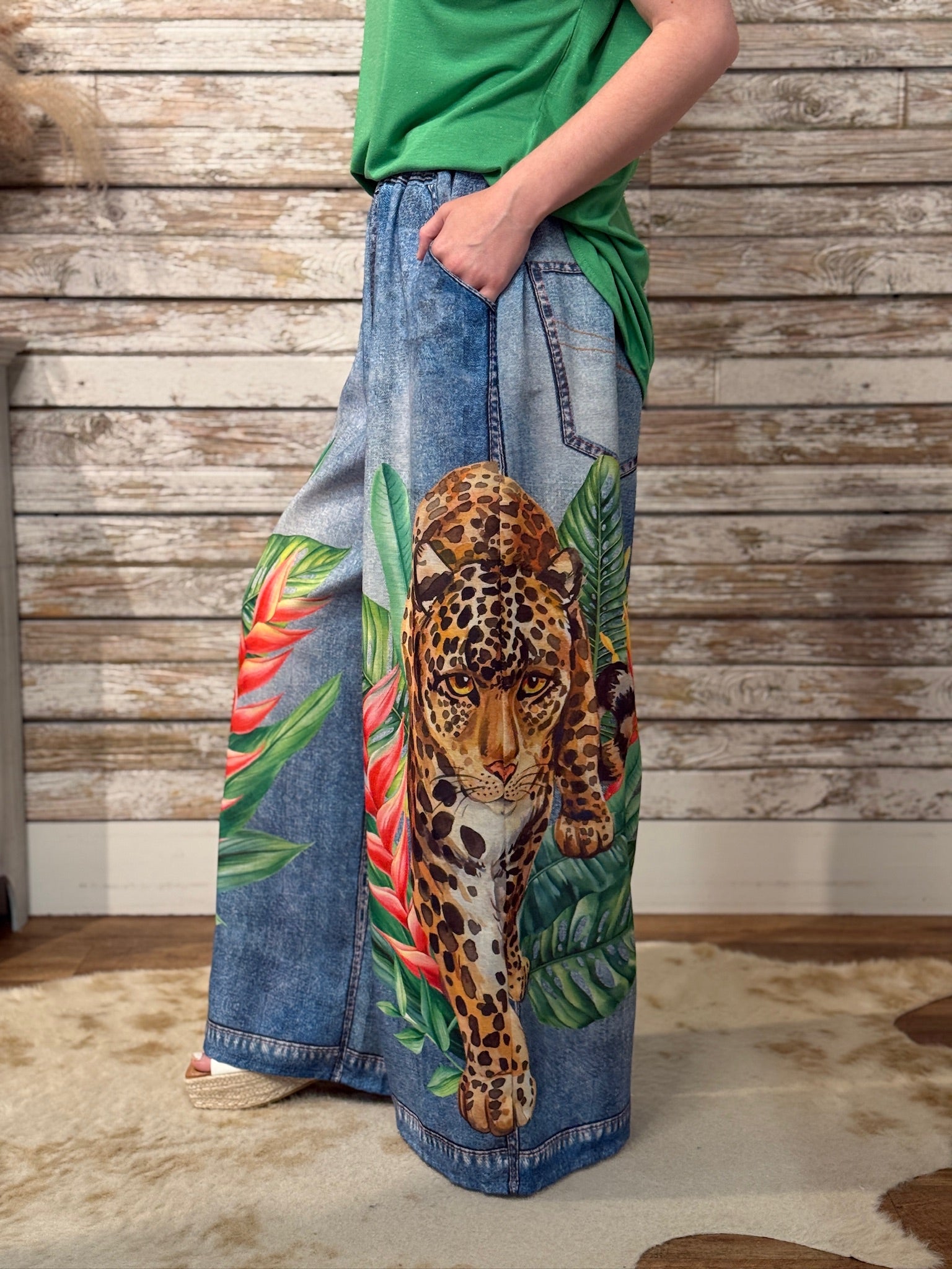 Person wearing a pair of wide-leg jeans with a leopard and tropical leaf design against a wooden wall.