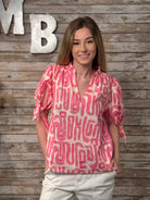 Woman wearing a pink patterned blouse standing against a wooden wall with decorative letters.
