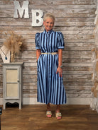 A woman modeling a navy and light blue vertically striped midi dress with puff sleeves and a gold belt, standing in front of a rustic wood-paneled wall with decorative letters and a small cabinet.