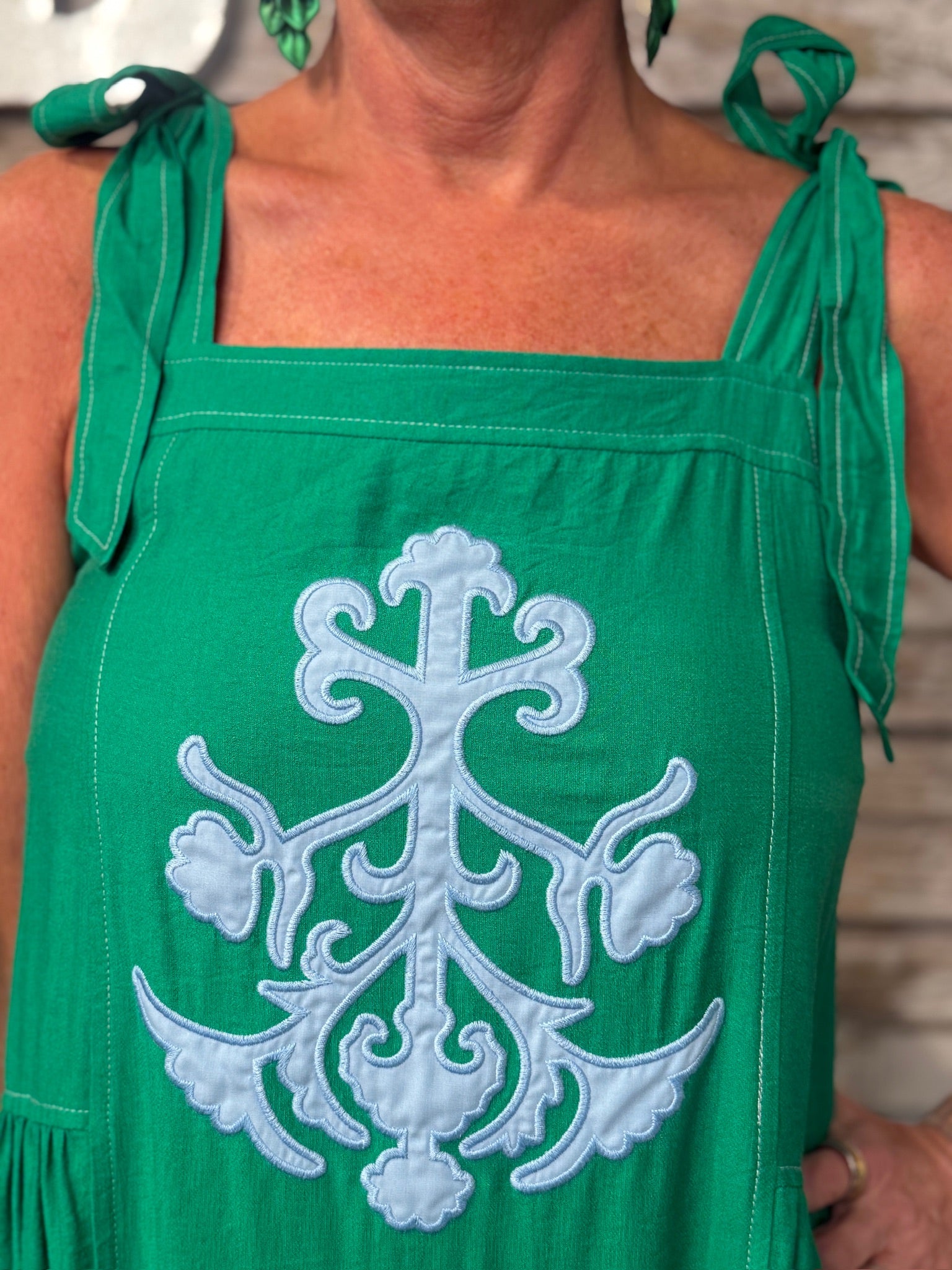 A close-up of a green sleeveless top or dress with a large light blue embroidered ornamental motif on the front and tie straps at the shoulders, worn by a person.