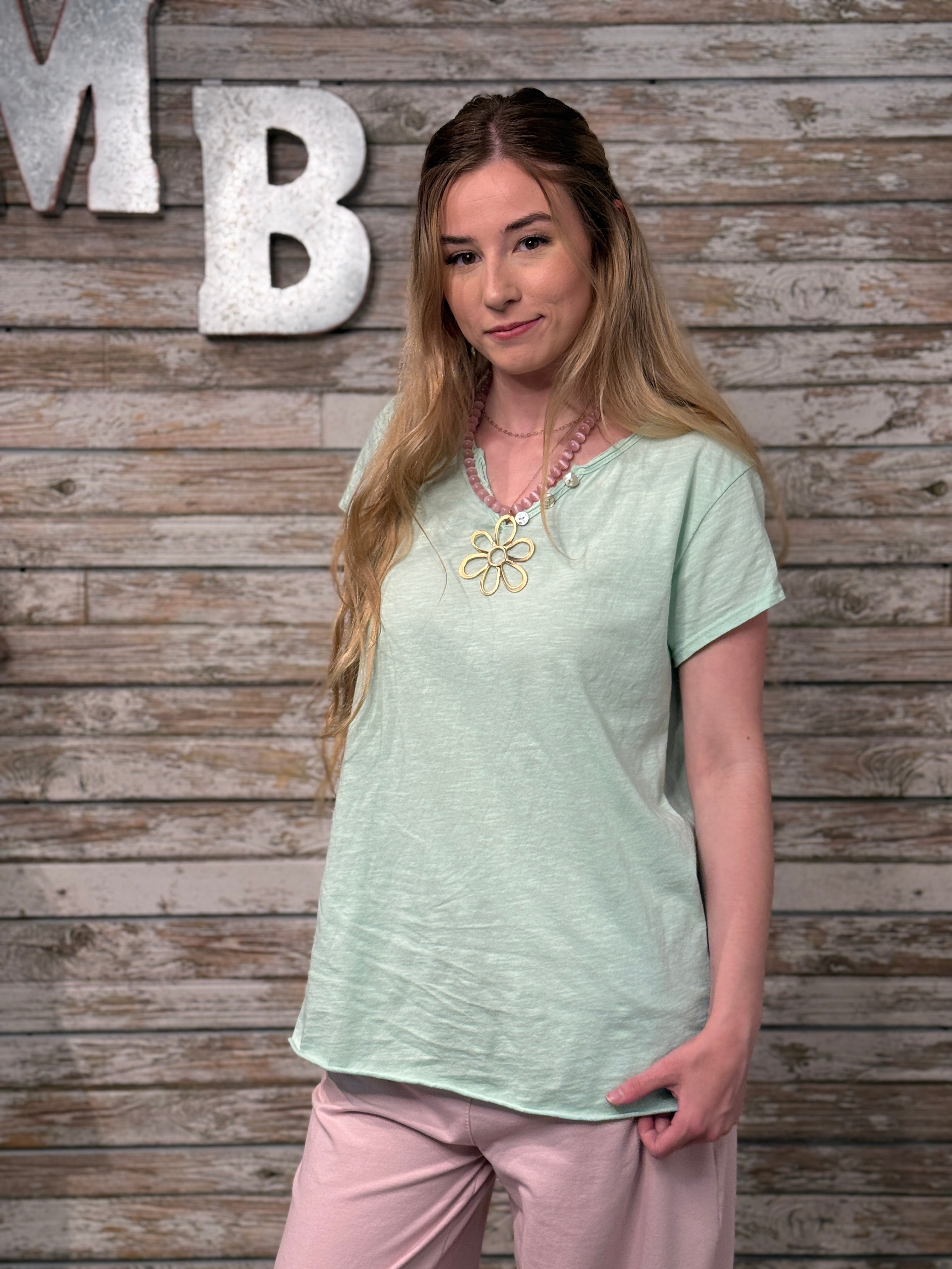 Woman wearing a light green top with a floral neckline and pink pants against a wooden wall.