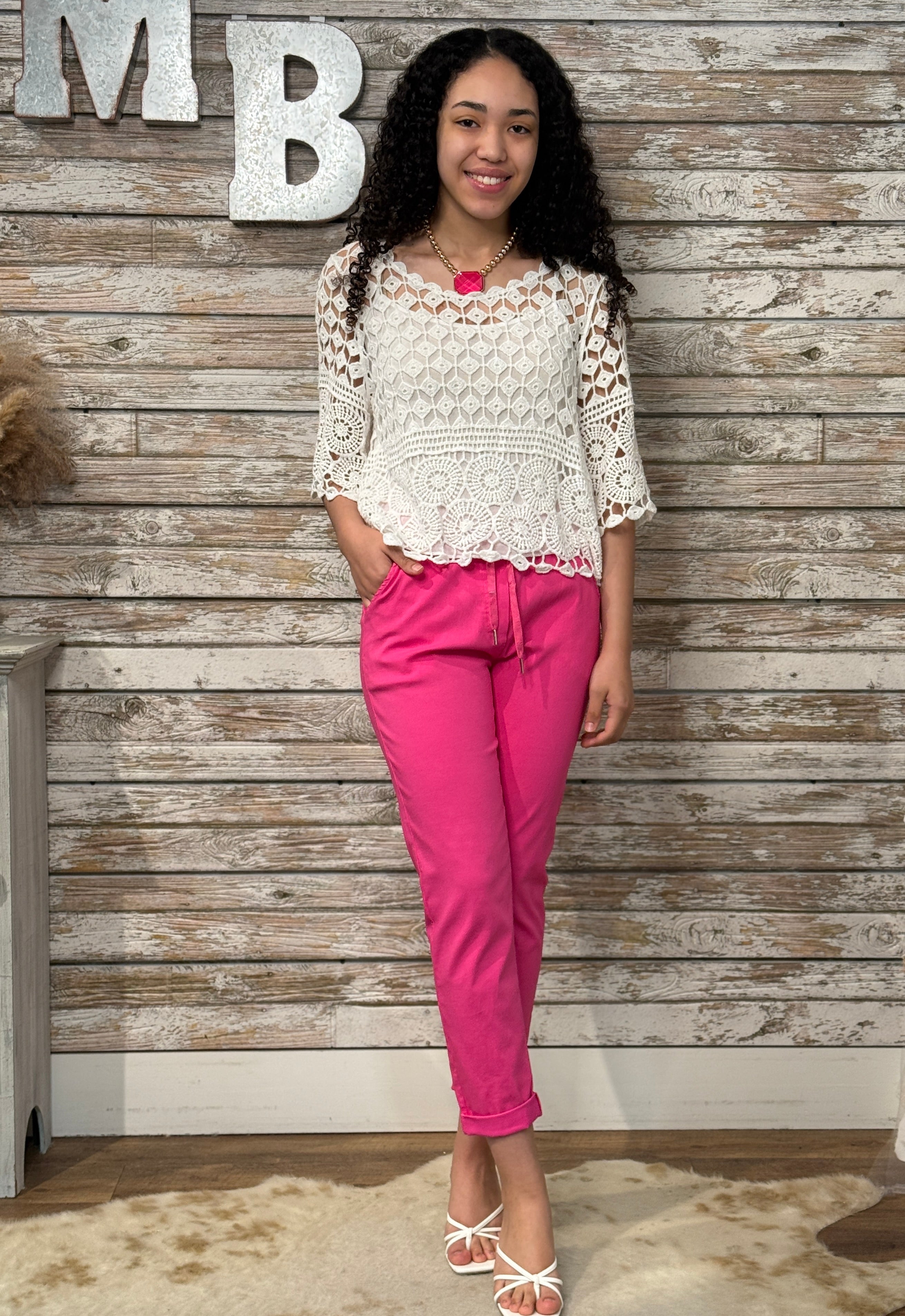 Woman in a white lace top and pink pants standing against a wooden paneled wall.