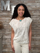 Woman wearing a white lace top and beige pants against a wooden wall with decorative letters.